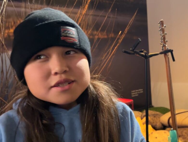 A girl with long dark hair and a black toque sits beside an instrument stand, and looks off to the side with a half-smile on her face.