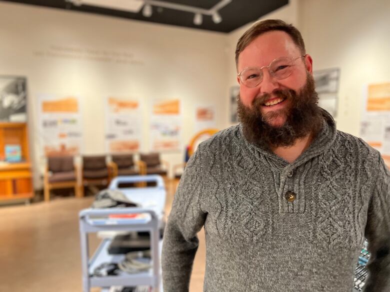 A large man wearing a grey knit sweater with a big beard and glasses smiles for the camera.