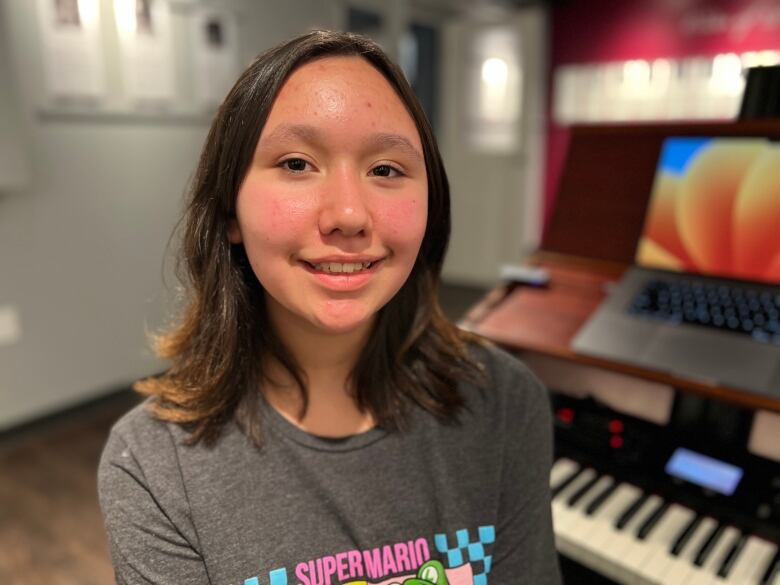 A young girl with long dark hair, wearing a Super Mario T-shirt, sits in front of a keyboard.