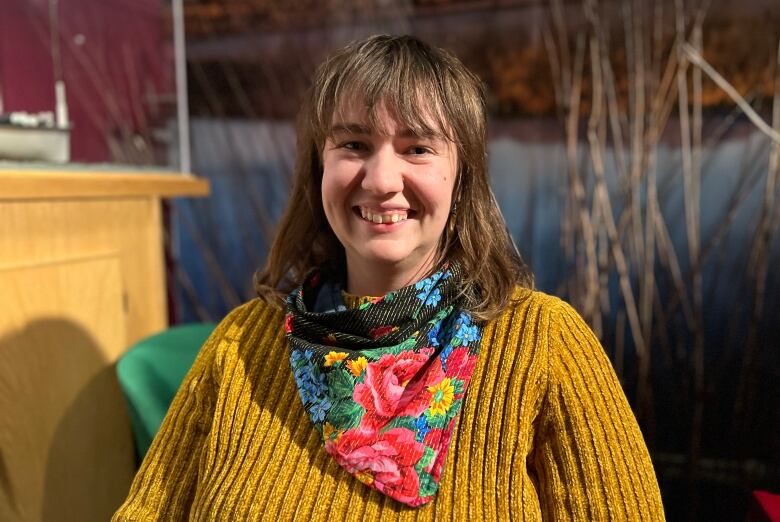 A woman with a yellow knit sweater and strawberry blond hair and a flowery scarf smiles for the camera.