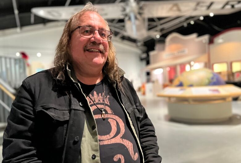 A man with long grey hair and a black jean jacket stands and smiles for the camera, the backdrop behind him is an airplane on display in a museum.