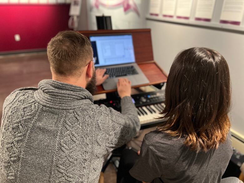 A man and a child sit next to eachother in front of a keyboard and computer. The camera photo is from behind them, looking over their shoulder.