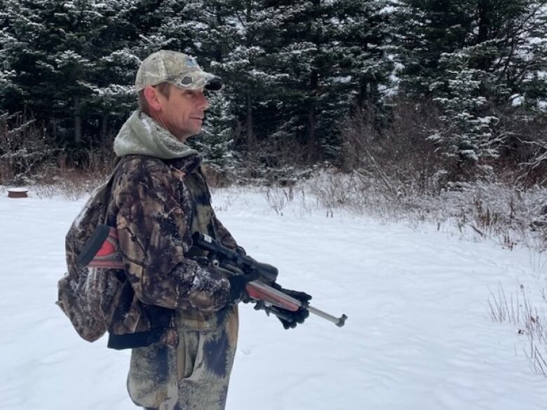 A man holding a hunting rifle standing in a wooded area covered with snow.