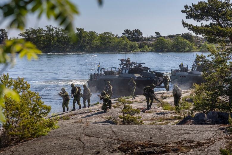 Armed soldiers in full gear advance on a rocky shoreline with two armed boats in the water behind them.