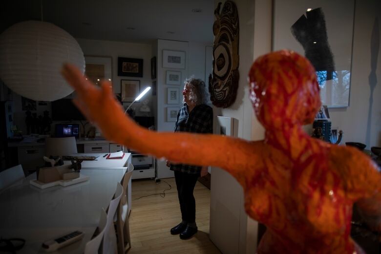 Susanna Blunt leans against a wall in the background behind a sculpture of a woman, painted in orange and red flames.