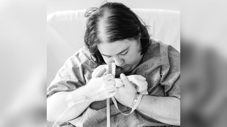 A black and white photo shows a woman in a hospital gown holding a tiny baby in a blanket. 