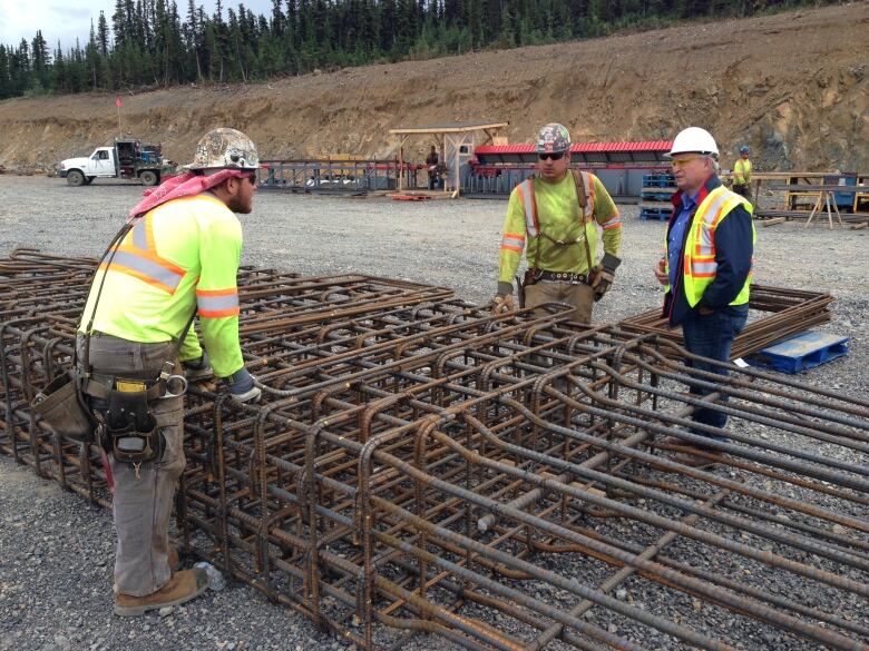 Three men in high-vis vests chat over iron struts in an outdoor environment.