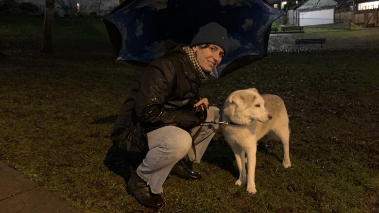 A woman holding an umbrella in rainy weather crouches in a park close to a dog which looks away from the camera.