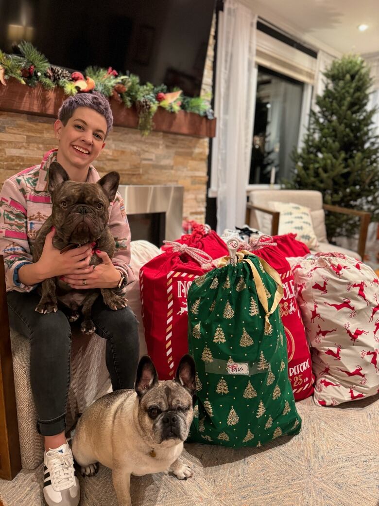 A woman sitting near a fire place holding a dog while another dog sits in front. 