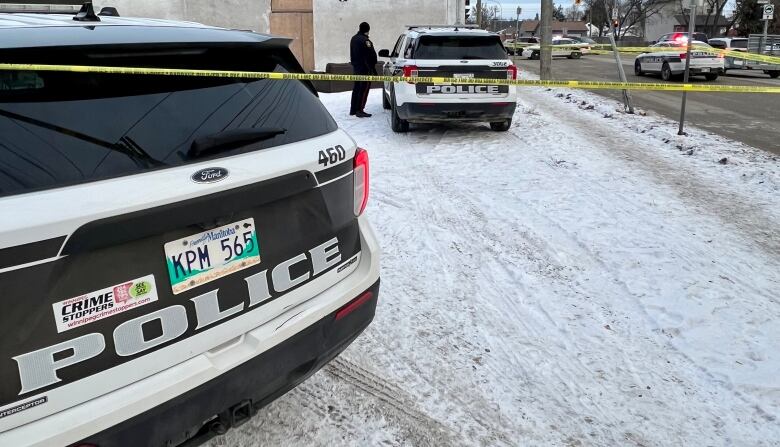 Police cars are parked in a snowy lot. Yellow police tape is stretched across a roadway. 