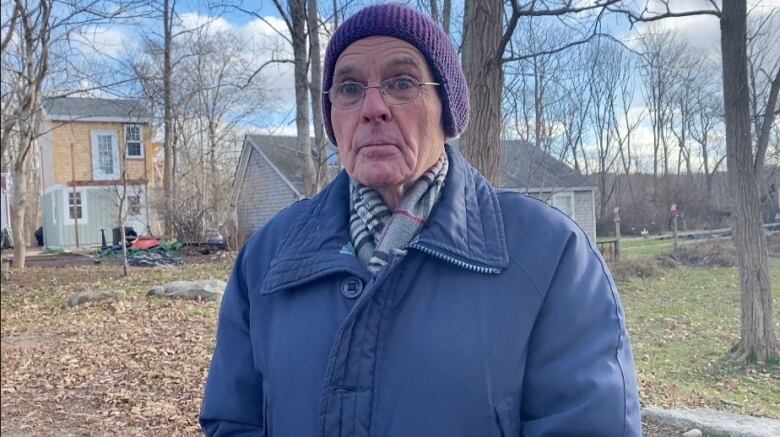 A white man with glasses and a purple knit toque and winter jacket stands outside with trees behind him, the new two-storey cabin in the background