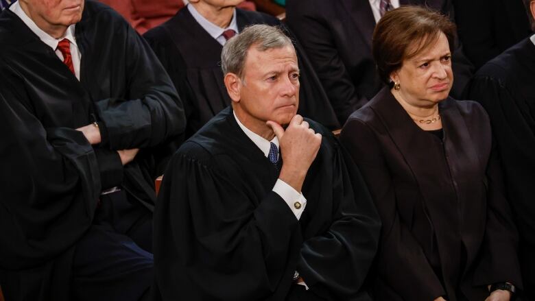 A cleanshaven man in a robe over top a collared shirt and tie sits his hand on his chin as a woman wearing black sits beside him.