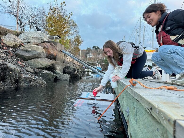 Two students lean off the side of a dock and place a baster into a hole of a plastic box that floats in the water.