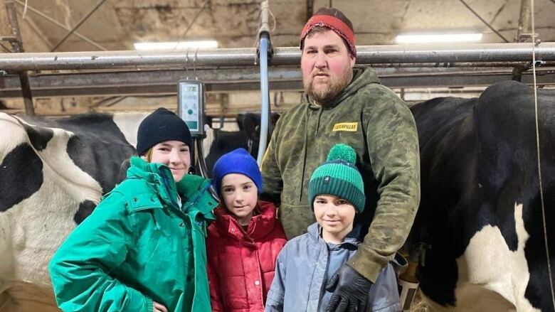 A man stands with his arms around his kids, standing in a barn with cows. 