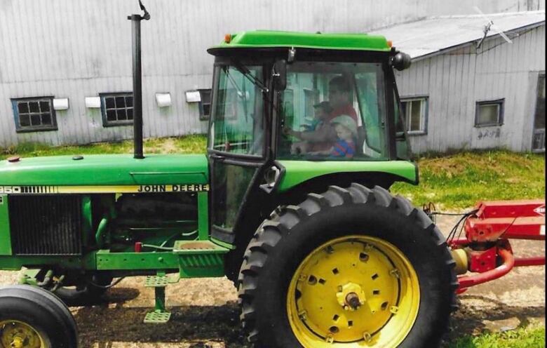 A green tracker on a farm. Inside the trackor a man is at the wheel, next to him are two kids. 