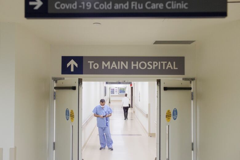 A healthcare worker wearing blue scrubs stands in a hallway under a sign that says 'To Main Hospital'