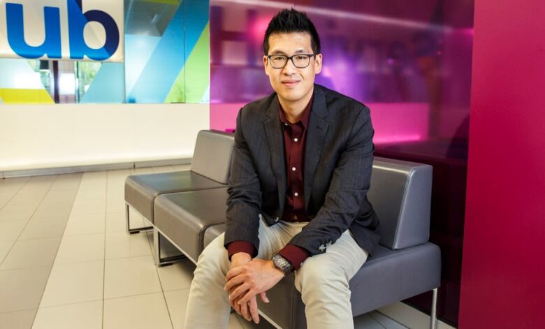 a man wearing glasses and a suit is sitting on a grey couch, smiling