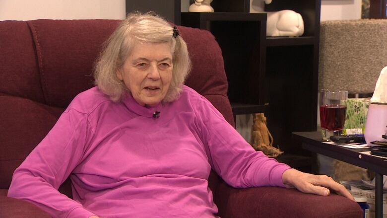 An elderly woman in a pink shirt sits in an armchair. 