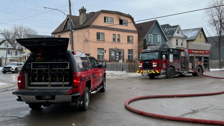 Fire trucks and fire hoses are in a street intersection while a building in the background is scorched from a fire.