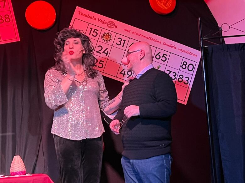 Two people on a stage, one wearing a curly brown wig and sequin top. 