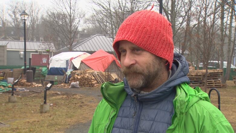 Mark wears an orange hat, green coat, and is standing in front of Tent City - the tent encampment at Bannerman Park in St. John's. 