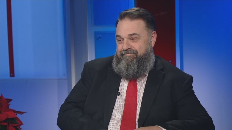 A man with a beard, dark suit and red tie sits in a TV studio. 