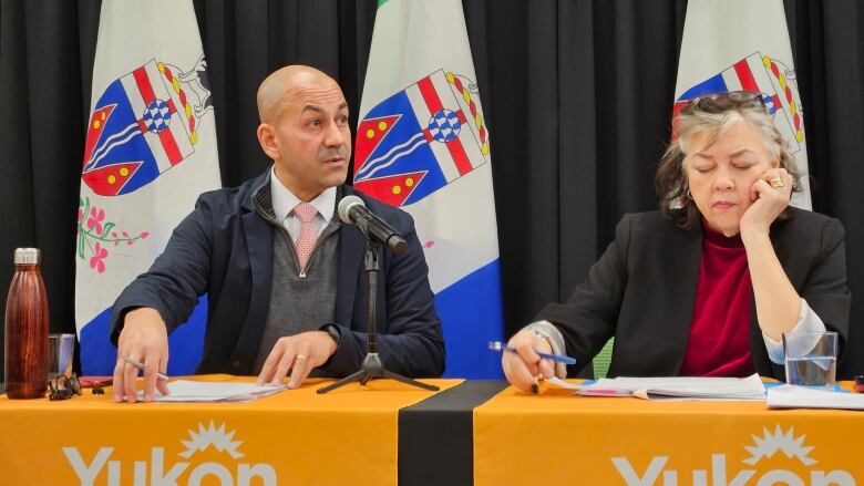 A man and a woman sit behind a table and microphone, in front of some flags.