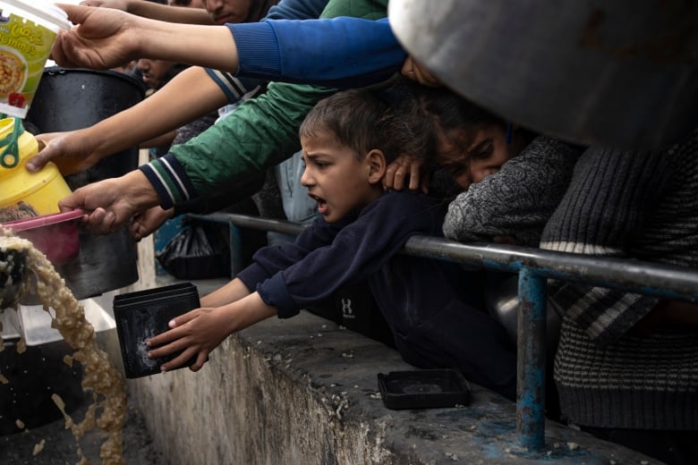 A young child standing with a crowd holds out a container for food