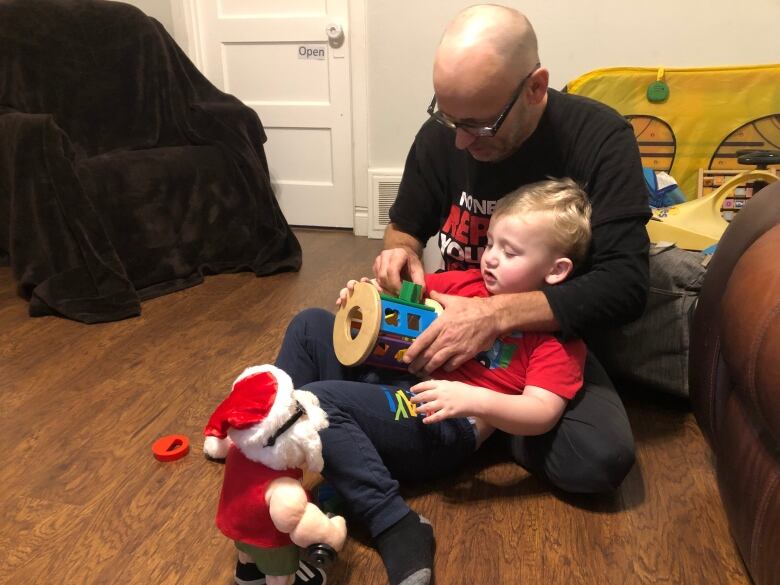 A young boy wearing a red t-shirt sitting in the lap of his bald father wearing a black shirt