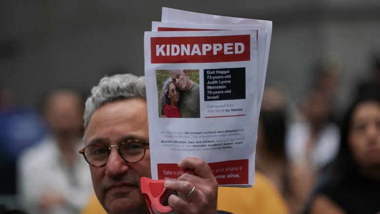 A bespectacled, clean shaven man holds up a paper that says, 'Kidnapped,' and contains a picture of an older man and woman. 