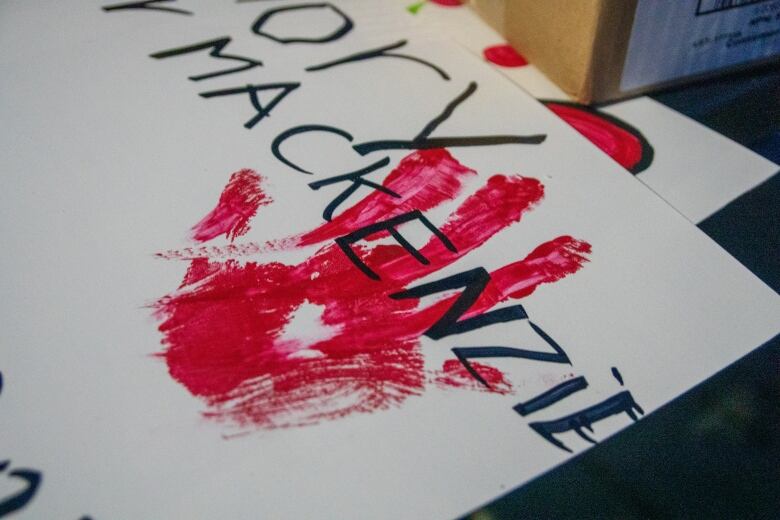 A red handprint is seen stamped over the handwritten name Mackenzie on a white poster.