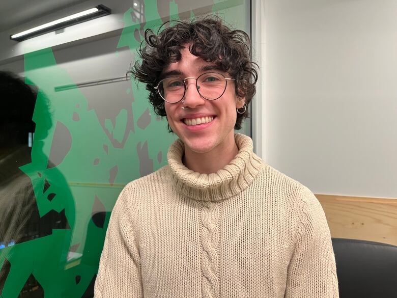 man with curly brown hair wearing a beige sweater with round glasses and piercings smiling at the camera.