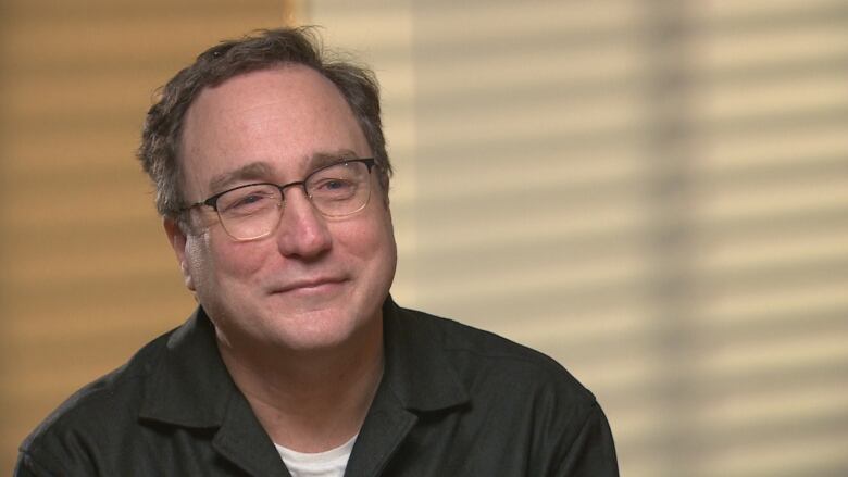 A man with glasses has a slight smile, wearing a black shirt against a plain wall. 