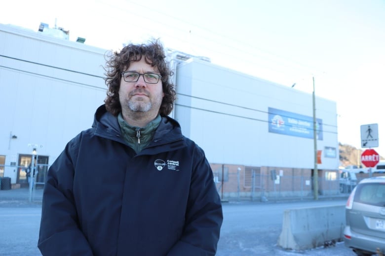 A man stands outside wearing a jacket. he has his back facing a building. 