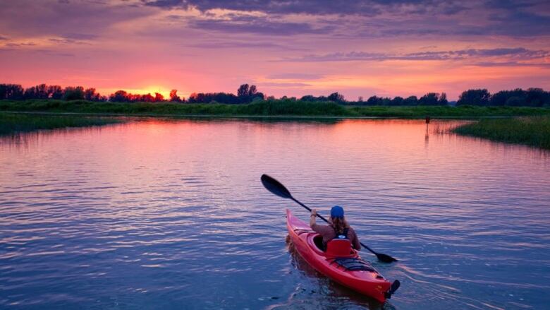 A woman kayaks on a river with a bright pink sunset in the background.