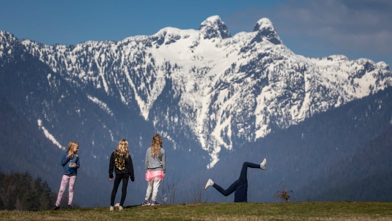 Three girls watching another kid doing a cartwheel on grass, overlooking snow-covered mountains.