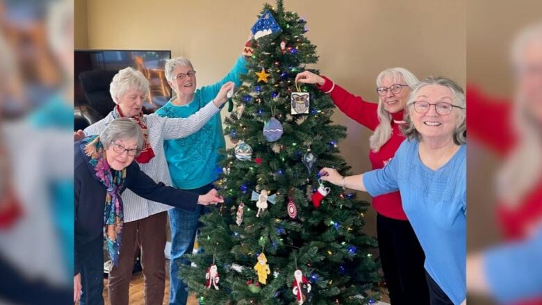 five women hold ornaments on an artificial Christmas tree and smile at a camera. 