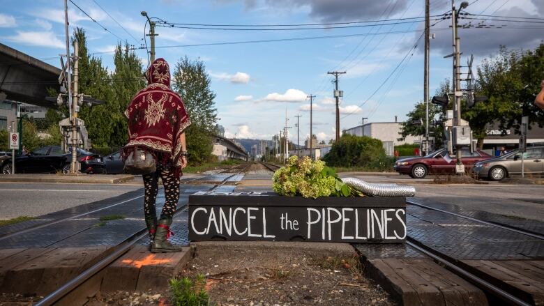 A person wearing a colourful red sweater stands on a railway line, with the words 'Cancel the Pipelines' visible on a black box below them.