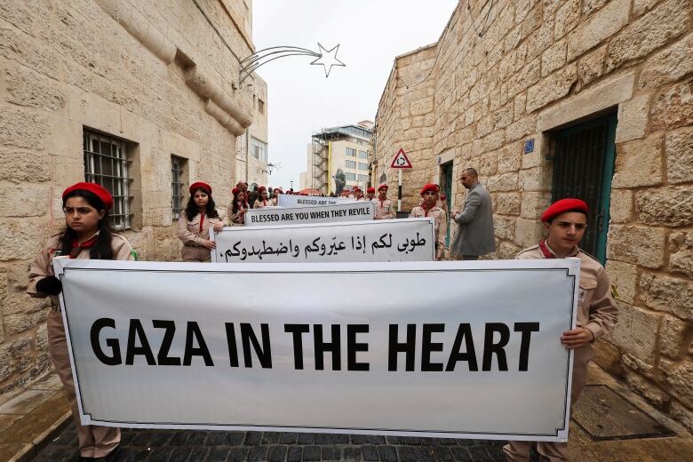 Children in an alley hold signs in English and Arabic. English sings say 'Gaza in the heart' and 'Blessed are you when they revile'.