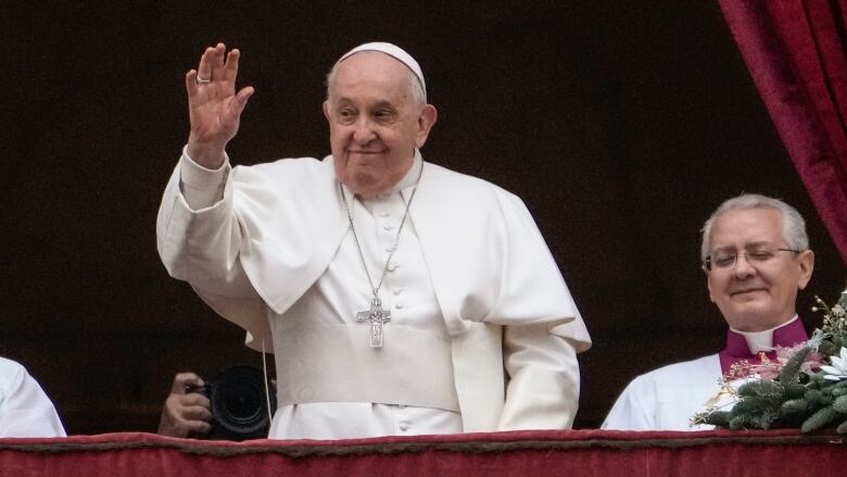 A man in white robes waves from a balcony. Another man stands next to him, smiling.