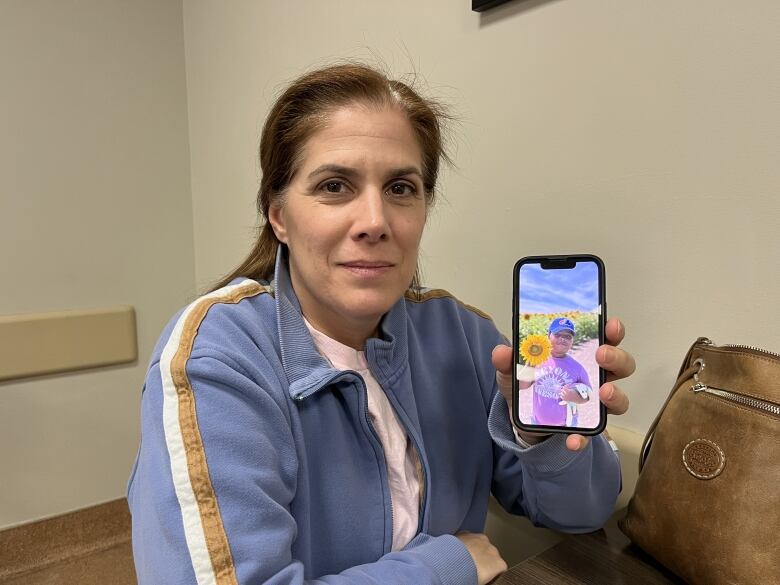 A woman sits at a table and holds up her phone showing a picture of a young boy. 