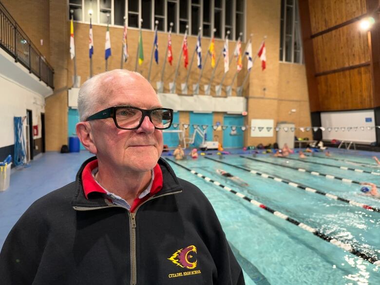 A man wearing glasses stands with a swimming pool shown in the background. 