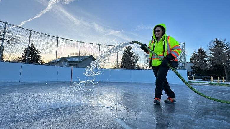 A woman in a high-viz jacket floods an outdoor rink with a hose of water.