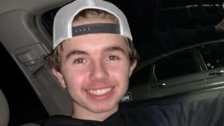 A young boy in a backwards baseball cap and a grey sweater sits in a vehicle.