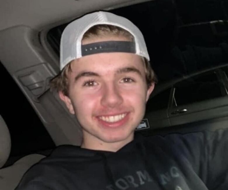 A young boy in a backwards baseball cap and a grey sweater sits in a vehicle.