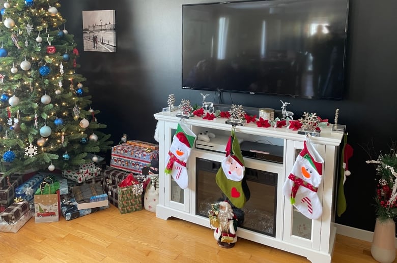 A Christmas tree with gifts underneath and a white fireplace mantle with three stockings in a family room.