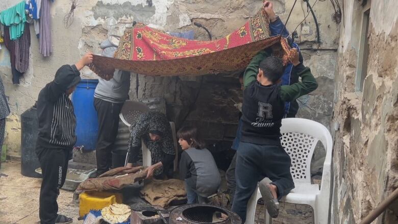 Four children hold the corners of a carpet above their heads, as a woman preparing food and a smaller child huddle beneath the carpet