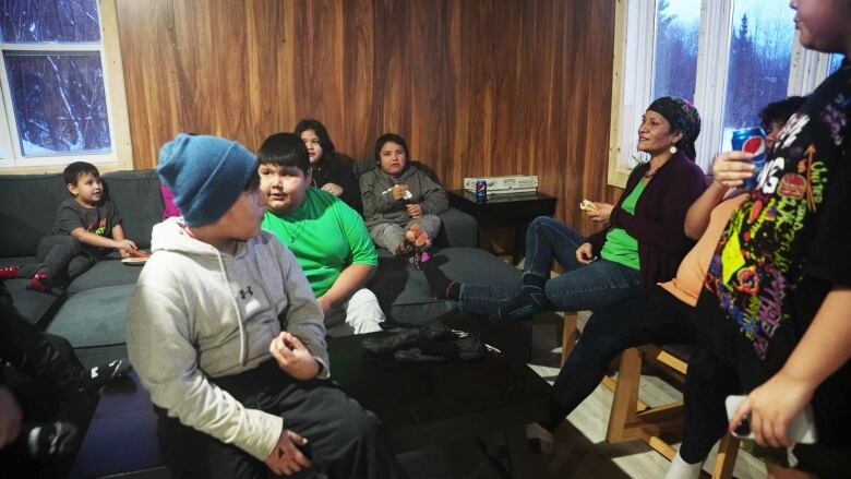 A group of kids sit around a large couch with one woman wearing a floral scarf sitting on the side. 