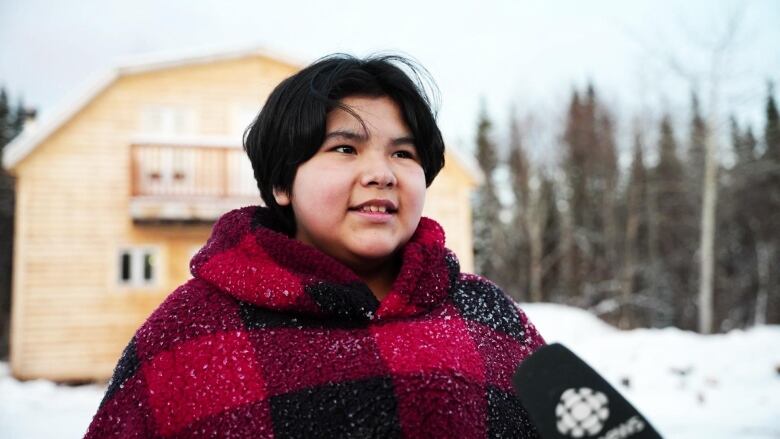 A young girl in a plaid sweater stands in front of a wooden cabin. 