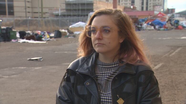 A person with dyed long pink hair and big glasses wearing a black leather jacket and standing in front of homeless encampments behind her. 